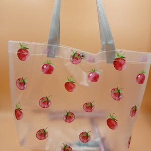 May include: A clear, semi-transparent tote bag featuring a repeating pattern of red watercolor-style tomatoes with green stems. The bag includes two light gray fabric shoulder straps and a single silver-toned metal snap button closure at the top.