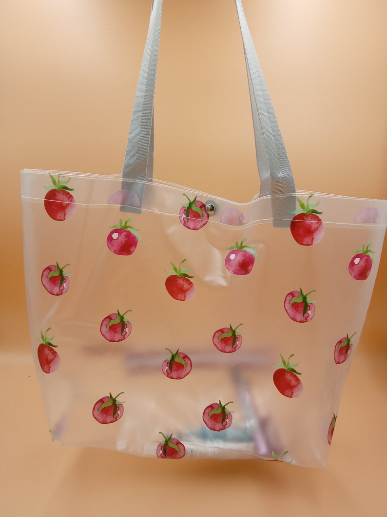 May include: A clear, semi-transparent tote bag featuring a repeating pattern of red watercolor-style tomatoes with green stems. The bag includes two light gray fabric shoulder straps and a single silver-toned metal snap button closure at the top.
