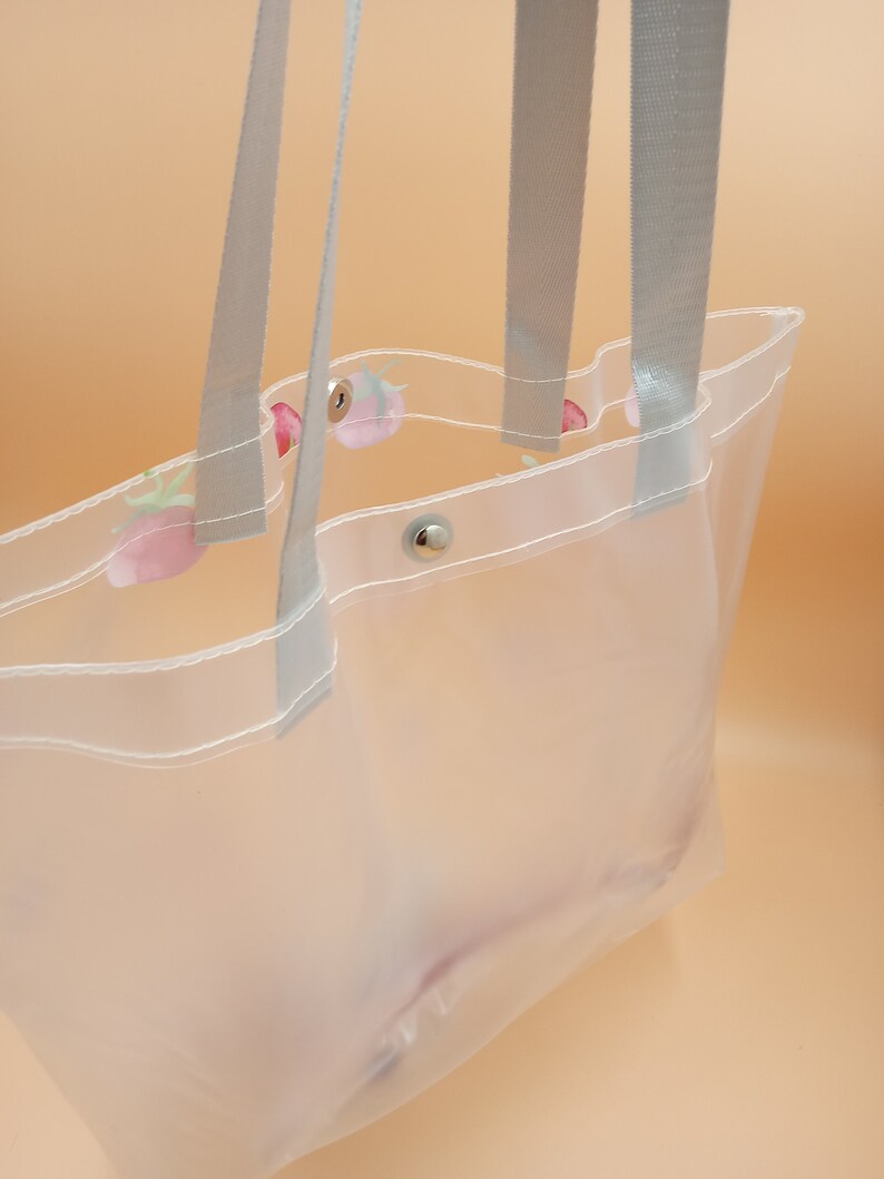 May include: A clear plastic tote bag featuring a repeating strawberry pattern along the top edge. The bag includes two light gray woven fabric handles and a single silver-toned metal snap button closure at the center of the opening.
