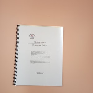 Op de afbeelding: Een wit spiraalgebonden naslagwerk getiteld 3D Organizer Reference Guide voor het 3D Embroidery System. De omslag bevat zwarte tekst, een logo en copyrightinformatie uit 2004, met een zwarte plastic spiraalbinding aan de linkerzijde.