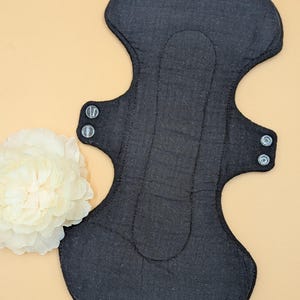 Puede incluir: Compresa menstrual de tela reutilizable negra con broches a los lados. La compresa está sobre una superficie marrón claro.