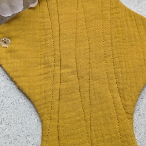 May include: A mustard yellow reusable cloth menstrual pad made of textured muslin fabric. The pad features a central stitched channel for absorbency and a clear plastic snap fastener on the side wing. A white peony flower is visible at the top.