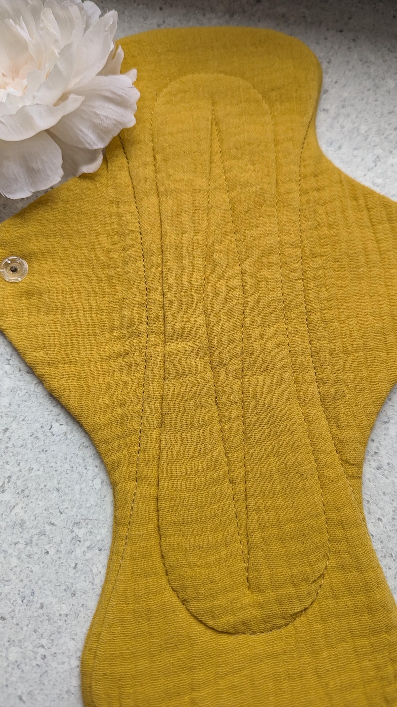 May include: A mustard yellow reusable cloth menstrual pad made of textured muslin fabric. The pad features a central stitched channel for absorbency and a clear plastic snap fastener on the side wing. A white peony flower is visible at the top.