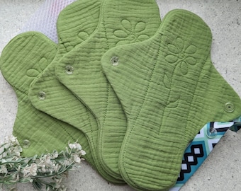 Könnte beinhalten: Ein Stapel aus sechs grünen wiederverwendbaren Stoff-Menstruationseinlagen mit Blumendesign. Die Einlagen sind gefaltet und fächerförmig angeordnet. Ein blaues und weißes Band ist um den unteren Teil des Stapels gewickelt.