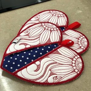 May include: Three red heart-shaped pot holders with white embroidered designs. The pot holders have a blue and white polka dot fabric on the back.