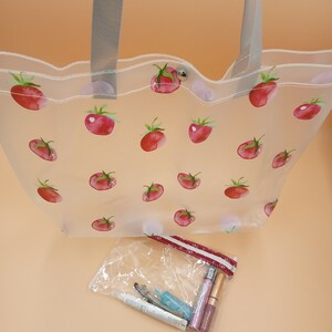 May include: A clear tote bag featuring a repeating pattern of red tomatoes with green stems, grey shoulder straps, and a silver snap closure. Below the bag is a small, clear zippered pouch containing several cosmetic tubes and applicators.