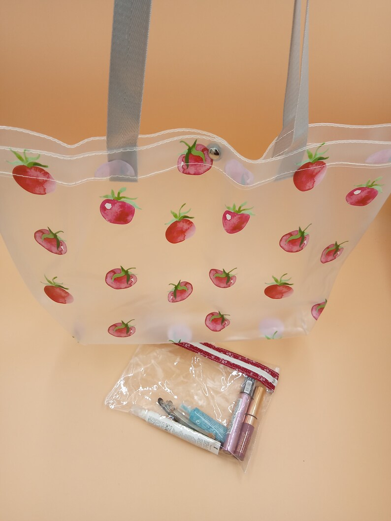 May include: A clear tote bag featuring a repeating pattern of red tomatoes with green stems, grey shoulder straps, and a silver snap closure. Below the bag is a small, clear zippered pouch containing several cosmetic tubes and applicators.