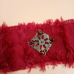 Puede incluir: Un brazalete de tela roja con un dije de filigrana plateado y cuentas marrones.