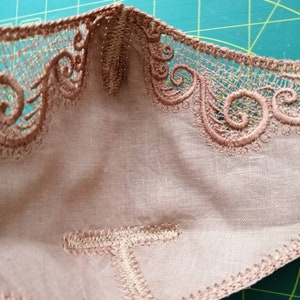May include: Close-up of the inside of a beige fabric mask with a brown lace trim. The mask is sewn with a zig-zag stitch.