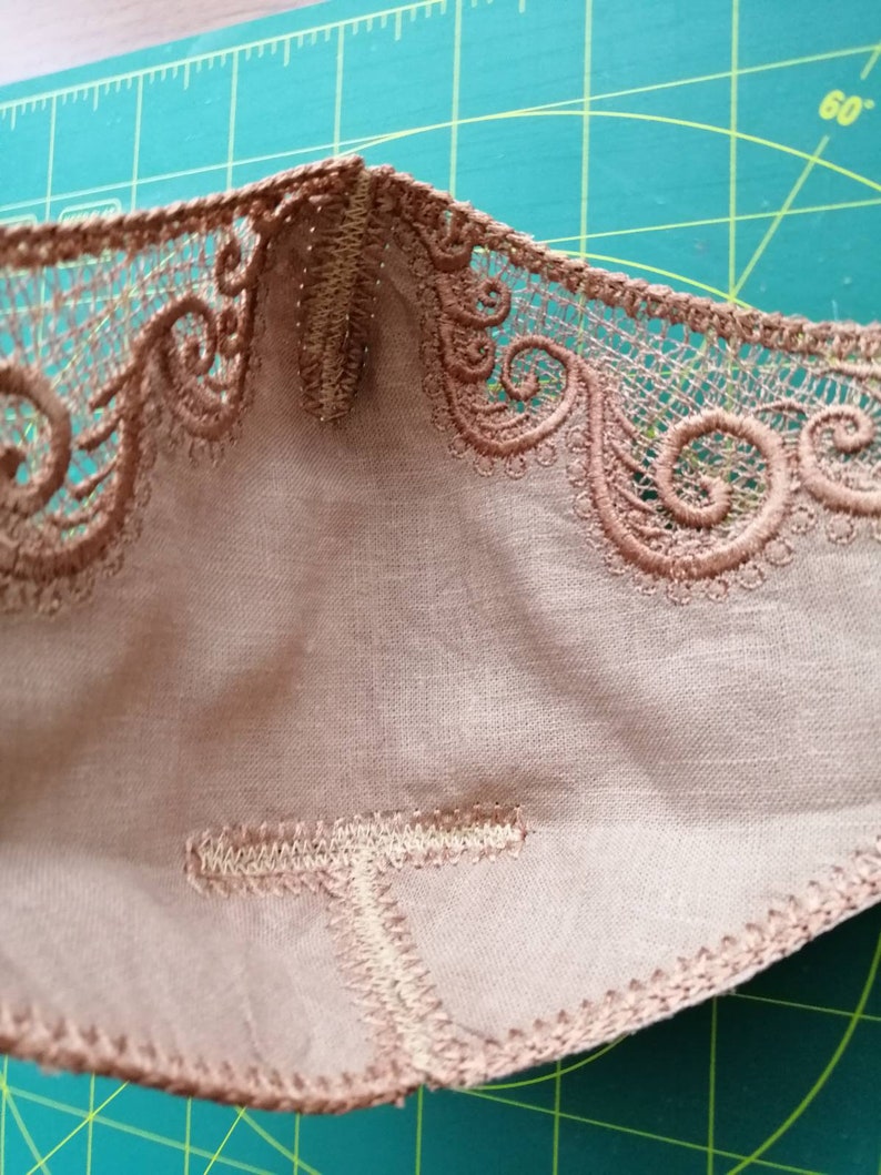 May include: Close-up of the inside of a beige fabric mask with a brown lace trim. The mask is sewn with a zig-zag stitch.