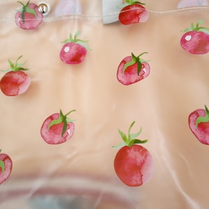 May include: A clear, semi-transparent tote bag featuring a repeating pattern of red watercolor-style tomatoes with green stems. The bag includes a light gray fabric strap at the top and a silver-toned metal snap button closure near the upper edge.