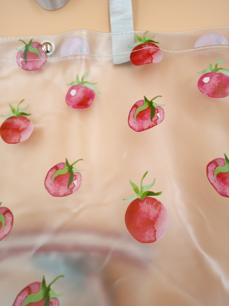 May include: A clear, semi-transparent tote bag featuring a repeating pattern of red watercolor-style tomatoes with green stems. The bag includes a light gray fabric strap at the top and a silver-toned metal snap button closure near the upper edge.