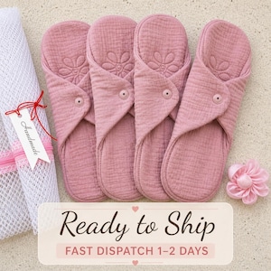 May include: Four reusable cloth menstrual pads in a dusty rose color, each featuring a quilted flower pattern and a single snap closure. A white mesh laundry bag with a tag labeled Handmade and a small pink fabric flower are positioned nearby.