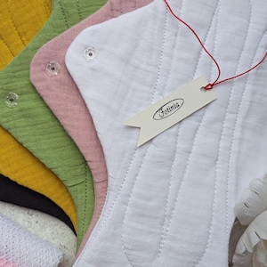 Può includere: Una pila di assorbenti in tessuto riutilizzabili nei colori bianco, rosa antico, verde salvia, giallo senape e nero. Ogni assorbente presenta una superficie in tessuto testurizzato, canali cuciti e bottoni a pressione trasparenti. È attaccata un'etichetta bianca con il testo Fotinia.