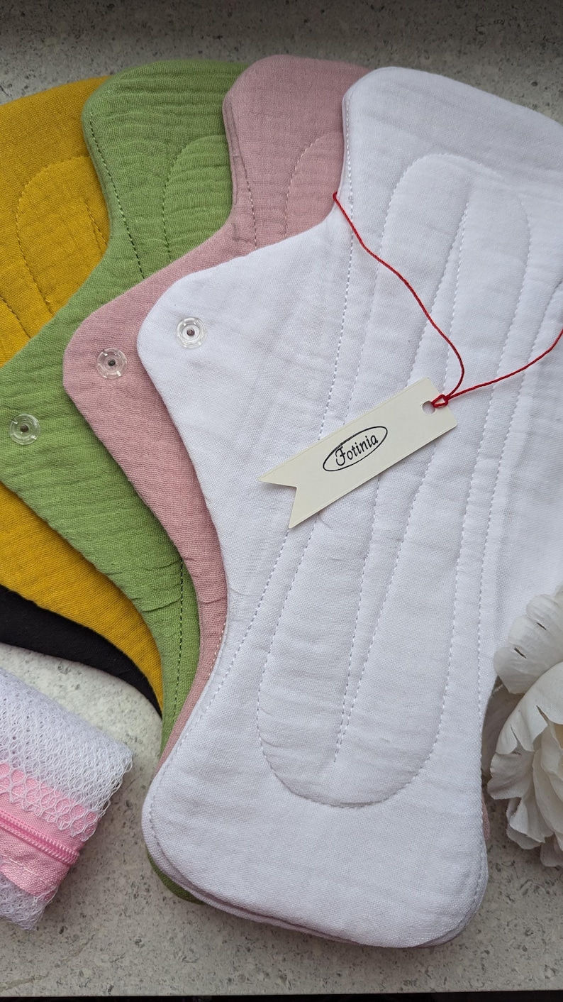 May include: A stack of reusable cloth pads in white, dusty pink, olive green, mustard yellow, and black. Each pad features a textured fabric surface, stitched channels, and clear plastic snap fasteners. A white tag with the text Fotinia is attached.