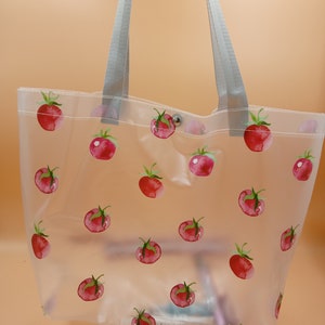 May include: A clear, semi-transparent tote bag featuring a repeating pattern of red watercolor-style tomatoes with green stems. The bag includes two light gray fabric shoulder straps and a single silver-toned metal snap button closure at the top.