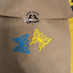 May include: A brown paper bag sealed with a sticker featuring a heart-shaped hand gesture and the text "STAY WITH Ukraine". Two die-cut butterfly ornaments, one blue and one yellow, are attached to the bag with thin silver strings.