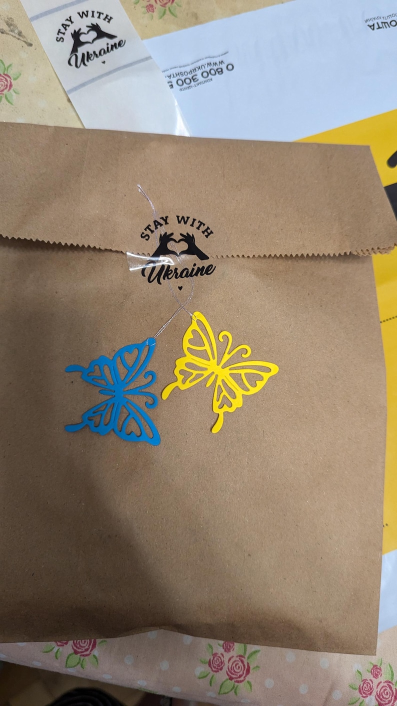 May include: A brown paper bag sealed with a sticker featuring a heart-shaped hand gesture and the text "STAY WITH Ukraine". Two die-cut butterfly ornaments, one blue and one yellow, are attached to the bag with thin silver strings.