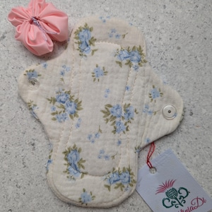 May include: A reusable cloth sanitary pad with a blue floral design on a cream background, secured with a snap. A pink fabric pouch and a tag with the "Folinia Di" logo are also visible.