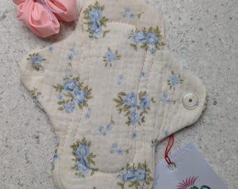 May include: A reusable cloth menstrual pad with a floral pattern in blue and cream, featuring a snap closure. A pink fabric pouch and a tag with a logo and the text "Folinia Di" are also present.