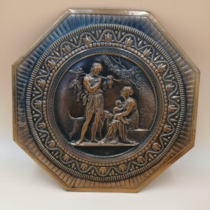 May include: Octagonal brass wall plaque with an embossed design of a family with two dogs. The plaque has a decorative border with a repeating pattern of stylized flowers.