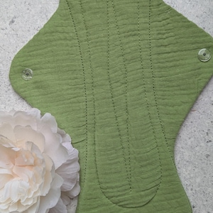 May include: A reusable cloth menstrual pad in a solid sage green color with a textured, crinkled fabric surface. The pad features a contoured shape with two clear plastic snap fasteners on the side wings and vertical stitched lines for structure.
