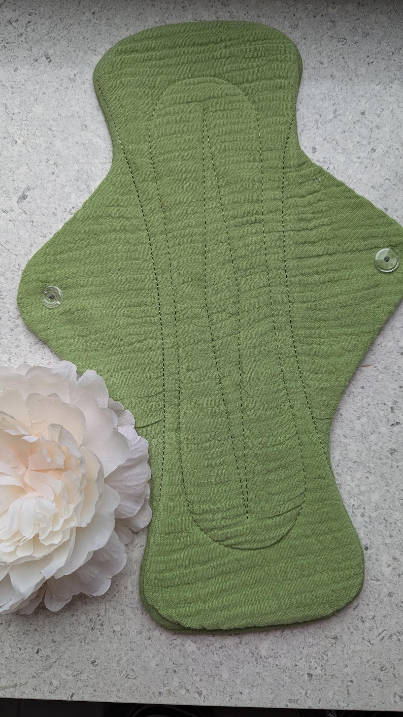 May include: A reusable cloth menstrual pad in a solid sage green color with a textured, crinkled fabric surface. The pad features a contoured shape with two clear plastic snap fasteners on the side wings and vertical stitched lines for structure.