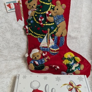 Op de afbeelding: Rode kerstsok van vilt met een witte viltboord. De sok is versierd met een groene kerstboom van vilt, drie bruine teddyberen, een blauwe zeilboot en een pop met geel haar. De sok heeft een label met de tekst "Prettige Kerst!"