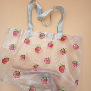 May include: A clear, semi-transparent tote bag featuring a repeating pattern of watercolor-style red tomatoes with green stems. The bag includes two light gray woven fabric handles and a single silver-toned metal snap button closure at the top center.