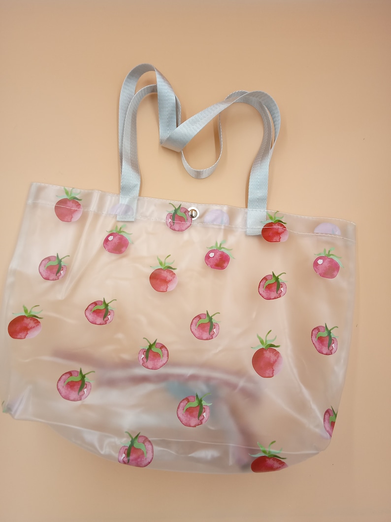 May include: A clear, semi-transparent tote bag featuring a repeating pattern of watercolor-style red tomatoes with green stems. The bag includes two light gray woven fabric handles and a single silver-toned metal snap button closure at the top center.