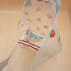 May include: A clear tote bag featuring a repeating pattern of red and pink strawberries. The bag has grey fabric handles, a silver snap button closure at the top, and an internal clear zippered pouch containing small cosmetic items.
