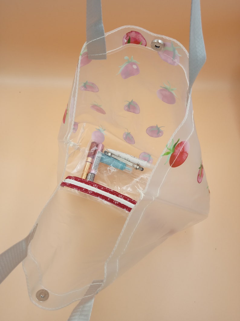 May include: A clear tote bag featuring a repeating pattern of red and pink strawberries. The bag has grey fabric handles, a silver snap button closure at the top, and an internal clear zippered pouch containing small cosmetic items.
