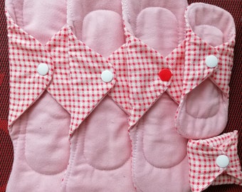 Könnte beinhalten: Vier wiederverwendbare Stoffbinden in Pink mit rotem und weißem Karomuster und weißen Druckknöpfen. Die Binden sind in absteigender Reihenfolge nach Größe angeordnet. Eine kleinere Karobinde befindet sich in der rechten unteren Ecke.