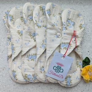 May include: Several reusable cloth menstrual pads with a cream background and blue floral pattern. Each pad is folded and secured with a button. A tag with the text "Fatinia Di" is attached.