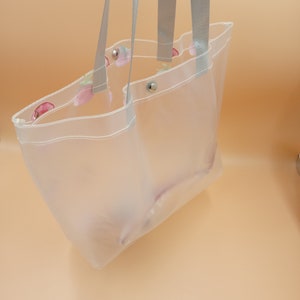 May include: A clear, translucent tote bag featuring a subtle strawberry pattern along the top edge. The bag includes two grey fabric shoulder straps and silver-toned metal snap button closures at the top opening for secure storage.