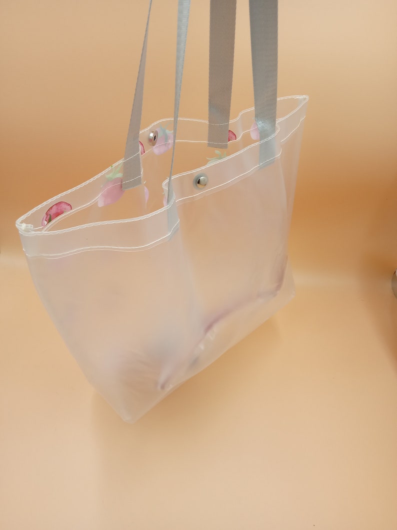 May include: A clear, translucent tote bag featuring a subtle strawberry pattern along the top edge. The bag includes two grey fabric shoulder straps and silver-toned metal snap button closures at the top opening for secure storage.