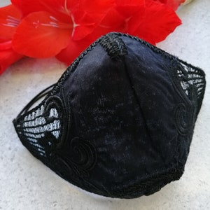 May include: A black face mask featuring a central seam and intricate black lace overlays on the sides. The mask has a structured, contoured shape with decorative embroidery details along the edges and thin black elastic ear loops for secure attachment.