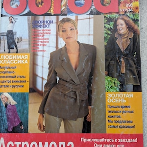 Könnte beinhalten: Ein Magazincover mit einer Frau, die einen braunen Blazer mit Gürtel trägt, und dem Text "Burda" in roten Buchstaben. Das Magazin heißt "AstroModa" und zeigt den Text "Goldener Herbst" in Gelb und Schwarz.