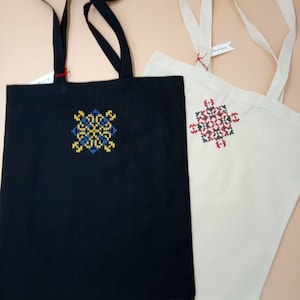 Può includere: Due borse tote, una nera e una bianco sporco, con disegni floreali ricamati. La borsa nera presenta un disegno giallo e blu, mentre la borsa bianco sporco ha un disegno rosso e nero. Entrambe le borse hanno manici lunghi e piccole etichette.