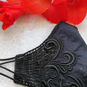 May include: A black face mask featuring intricate black embroidery with swirling patterns and a delicate lace side panel. The mask is constructed from solid black fabric and includes thin black elastic ear loops for secure attachment.