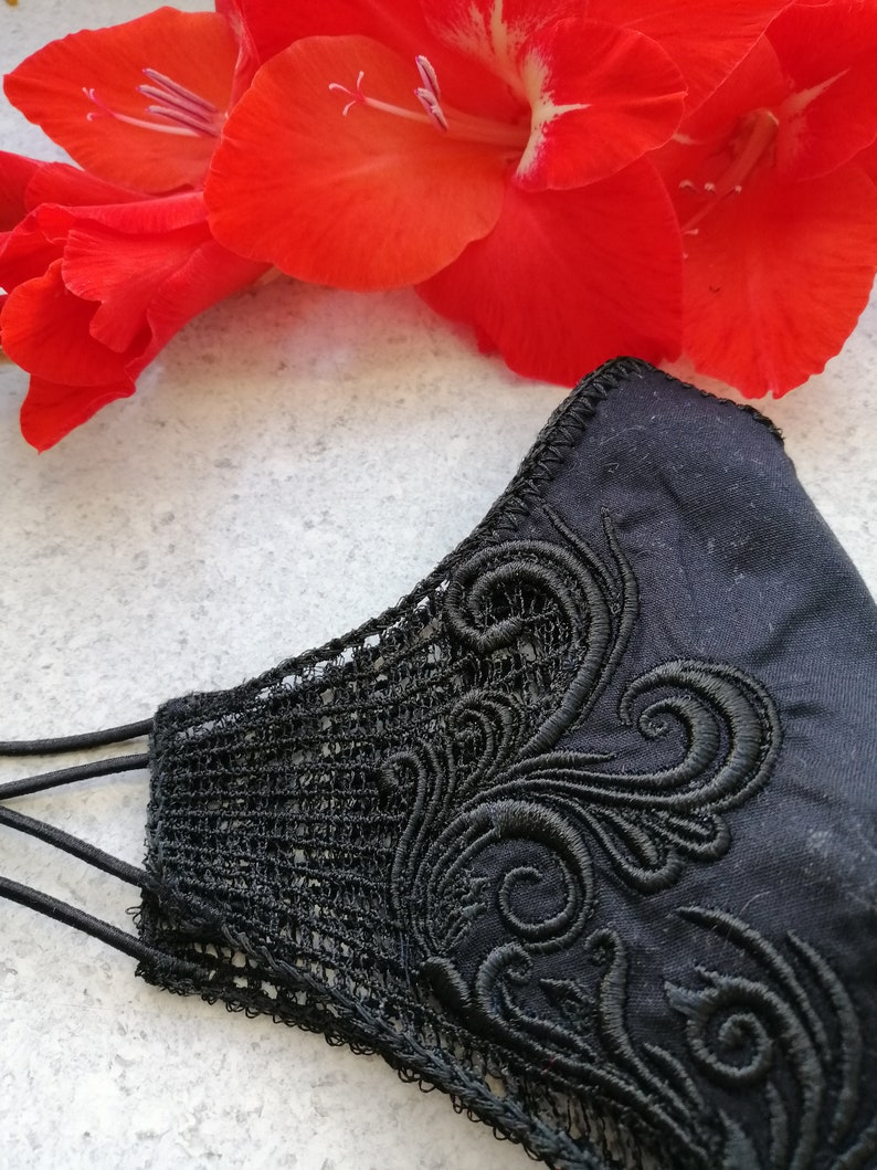 May include: A black face mask featuring intricate black embroidery with swirling patterns and a delicate lace side panel. The mask is constructed from solid black fabric and includes thin black elastic ear loops for secure attachment.