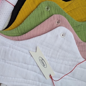 May include: A set of reusable cloth menstrual pads in white, dusty pink, sage green, mustard yellow, and black, featuring snap closures and a white tag with the text Fotinia. A small white mesh laundry bag with a pink zipper is included in the set.