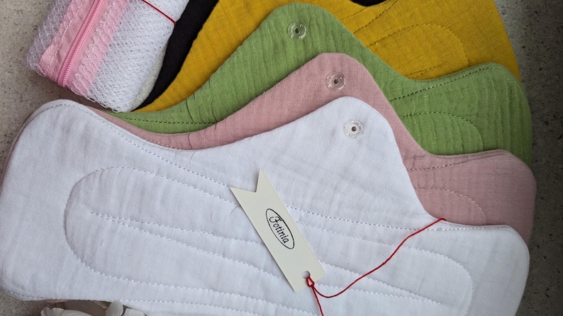 May include: A set of reusable cloth menstrual pads in white, dusty pink, sage green, mustard yellow, and black, featuring snap closures and a white tag with the text Fotinia. A small white mesh laundry bag with a pink zipper is included in the set.