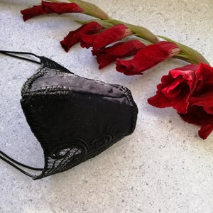 May include: A black face mask featuring a smooth central panel and intricate black lace trim along the edges, secured with thin black elastic ear loops. The mask is positioned next to a long stem of deep red gladiolus flowers with green leaves.