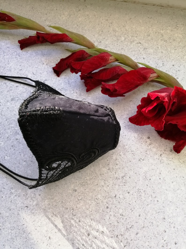 May include: A black face mask featuring a smooth central panel and intricate black lace trim along the edges, secured with thin black elastic ear loops. The mask is positioned next to a long stem of deep red gladiolus flowers with green leaves.