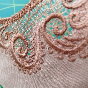 May include: Close-up of a beige fabric with a brown lace trim. The lace has a delicate, intricate design with a repeating pattern of swirls.
