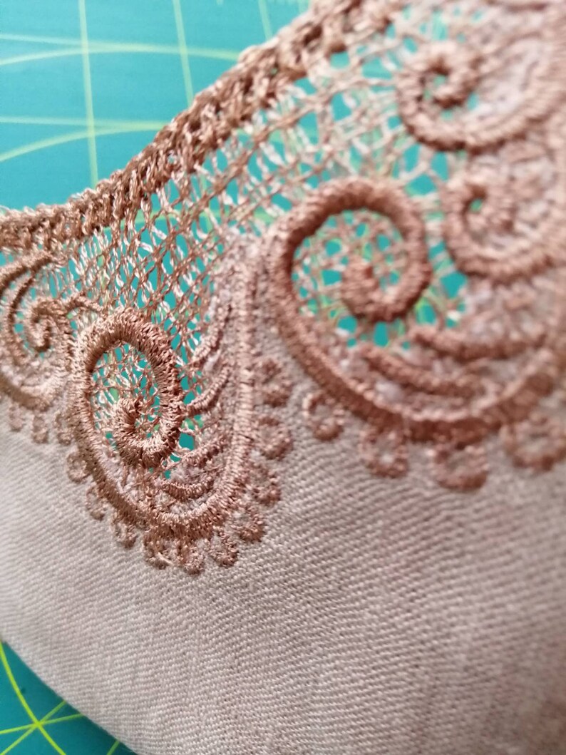 May include: Close-up of a beige fabric with a brown lace trim. The lace has a delicate, intricate design with a repeating pattern of swirls.