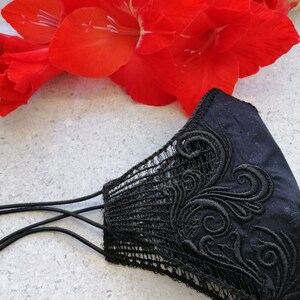 May include: A black face mask featuring intricate black embroidery with swirling patterns on the side and a delicate black lace trim along the edge. The mask includes thin black elastic ear loops for securing the item in place during use.