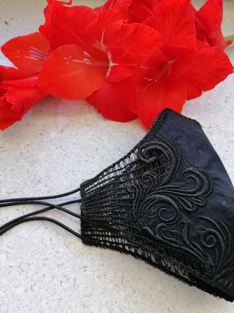 May include: A black face mask featuring intricate black embroidery with swirling patterns on the side and a delicate black lace trim along the edge. The mask includes thin black elastic ear loops for securing the item in place during use.