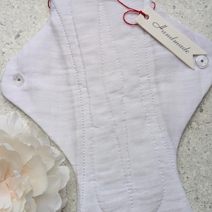 May include: A white cloth menstrual pad featuring vertical quilted stitching lines and two clear plastic snap buttons on the side wings. A small off-white tag with the word Handmade printed in black is attached by a thin red string.